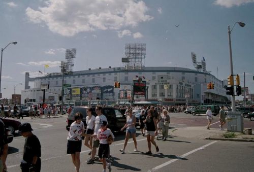 Crowd of people walking near a stadium on a sunny day, with cars and traffic lights visible in the foreground.