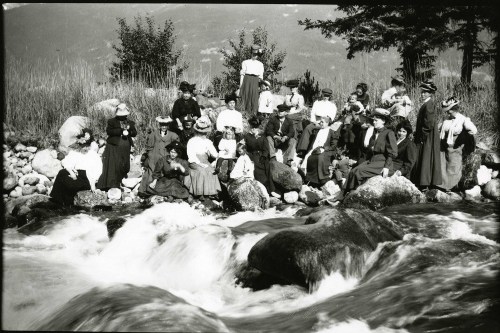 A large group of women in vintage clothing gathered by a rocky riverbank, surrounded by trees and mountains.