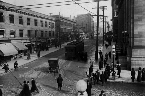Historic black-and-white street scene with a tram, early cars, and pedestrians in a bustling urban setting.