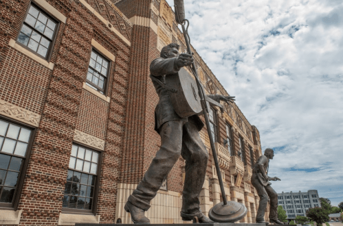 Two large bronze statues of musicians stand outside a brick building, one playing a guitar and the other holding a microphone.