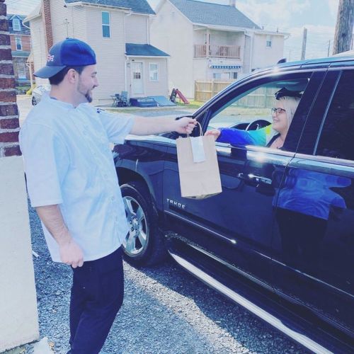 A chef hands a takeout bag to a woman in a car at a drive-thru.