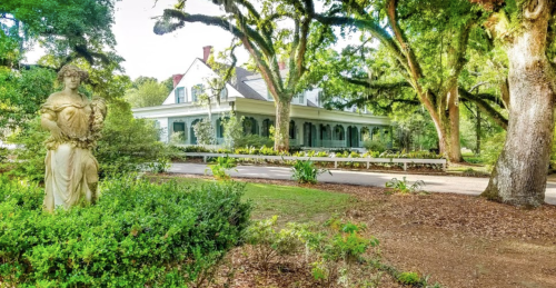 A historic house surrounded by lush greenery and trees, with a statue in the foreground.