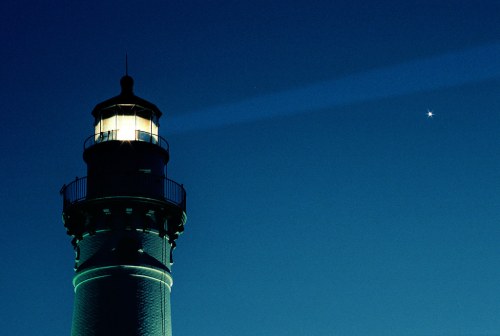 A lighthouse illuminated at night, with a bright star visible in the dark blue sky above.