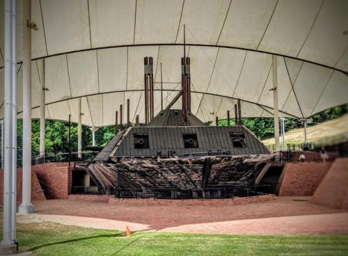 A historic ship displayed under a large canopy, surrounded by a grassy area and brick pathways.