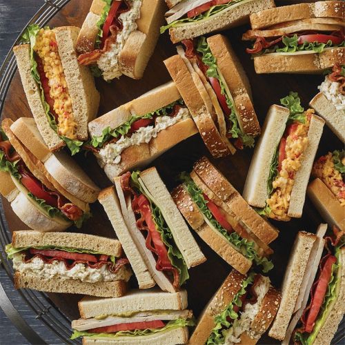 A variety of sandwiches stacked on a wooden platter, featuring lettuce, tomatoes, bacon, and different fillings.