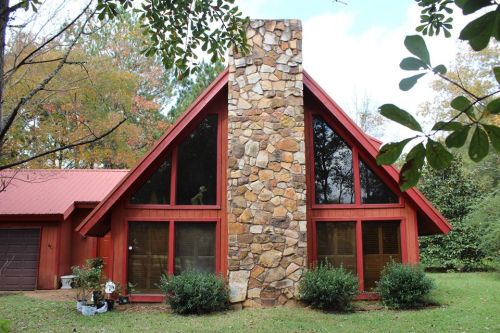A red A-frame house with a stone chimney, surrounded by greenery and trees in a serene outdoor setting.
