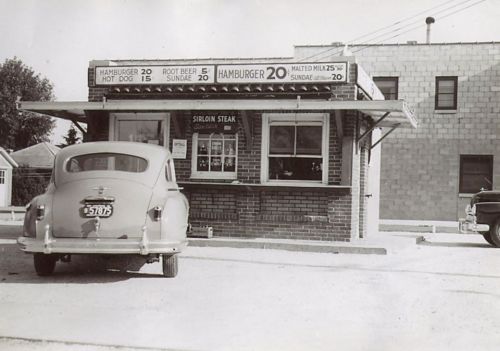 A vintage drive-in diner with a menu board, featuring burgers and hot dogs, and classic cars parked outside.