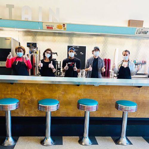 Five masked staff members stand behind a counter, giving thumbs up, with retro-style stools in front.