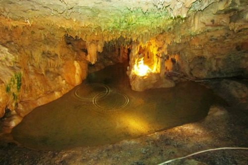 A dimly lit cave with a small pool of water reflecting light, surrounded by rocky formations and stalactites.