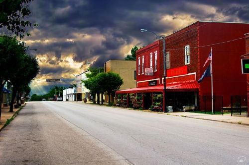 A quiet street scene with a red building, trees, and dramatic clouds overhead, creating a moody atmosphere.