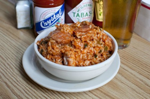 A bowl of flavorful rice mixed with sausage and spices, accompanied by bottles of hot sauce and a glass of beer.