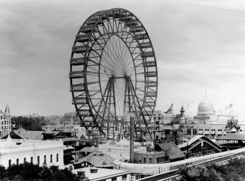 Historic black-and-white photo of a large Ferris wheel at an amusement park, surrounded by buildings and attractions.