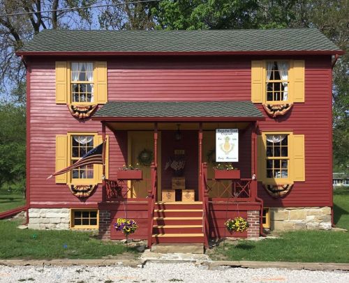 A charming red two-story house with yellow shutters, a porch, and flower pots, surrounded by green grass and trees.