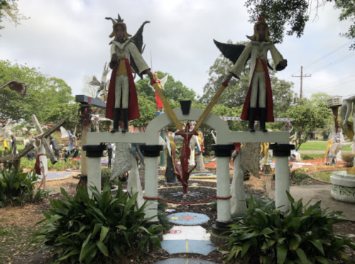 Two angelic statues with wings stand atop an archway in a vibrant outdoor setting, surrounded by greenery and decorations.
