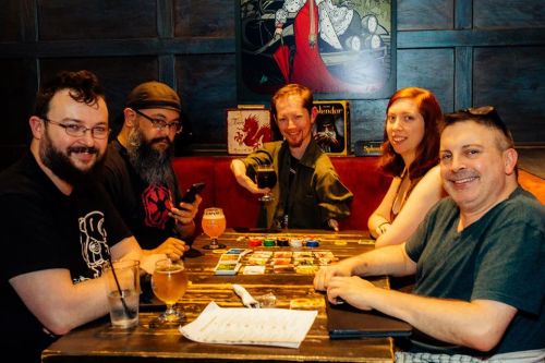 A group of five friends sits around a table with board games and drinks, smiling and enjoying their time together.