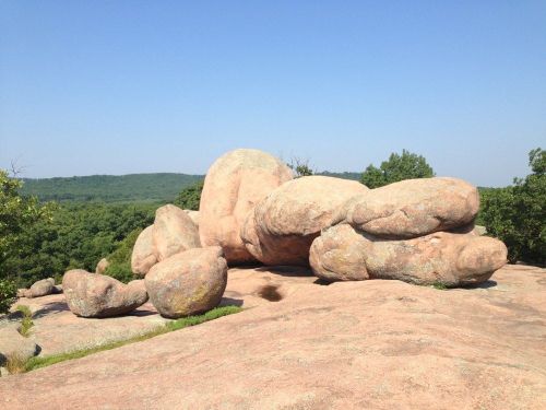 Large, smooth boulders scattered on a rocky surface, surrounded by green trees and a clear blue sky.