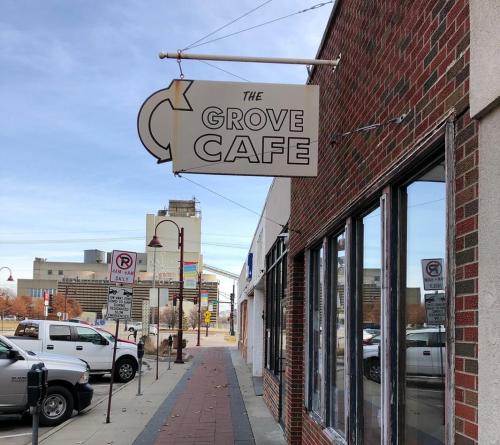 Sign for "The Grove Cafe" hanging above a brick building, with a view of a city street and parked cars.