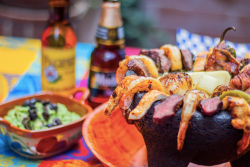 A vibrant table setting featuring a sizzling mixed grill with meat, vegetables, and drinks in the background.