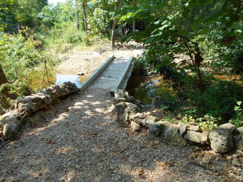 A wooden bridge spans a small stream, surrounded by lush greenery and rocky pathways in a serene forest setting.