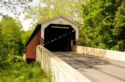 A red covered bridge with a white roof, surrounded by lush green trees, leading to a paved road.