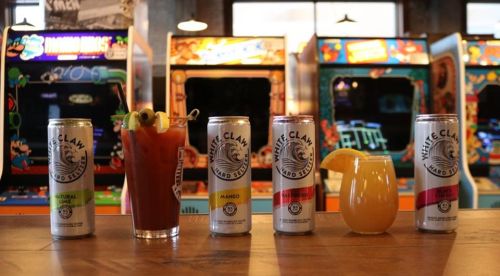 A bar scene featuring a cocktail and three cans of White Claw hard seltzer, with vintage arcade games in the background.