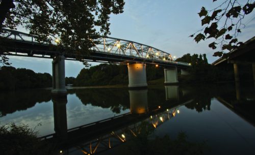 A bridge spans a calm river at dusk, with reflections in the water and soft lights illuminating its structure.