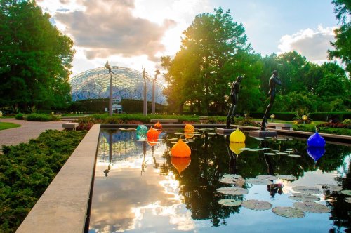 A serene garden scene with colorful glass sculptures in a pond, surrounded by lush greenery and a glass conservatory in the background.