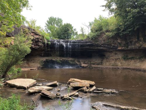 A serene waterfall cascades over rocky cliffs into a calm pool, surrounded by lush greenery and natural rock formations.