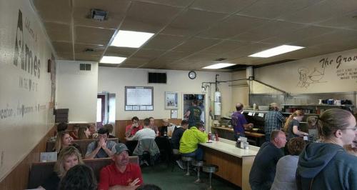 A busy diner interior with patrons seated at tables and a counter, and staff working in the kitchen area.