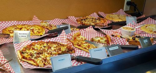 A variety of pizzas displayed on a counter, each labeled with toppings, on red and white checkered paper.