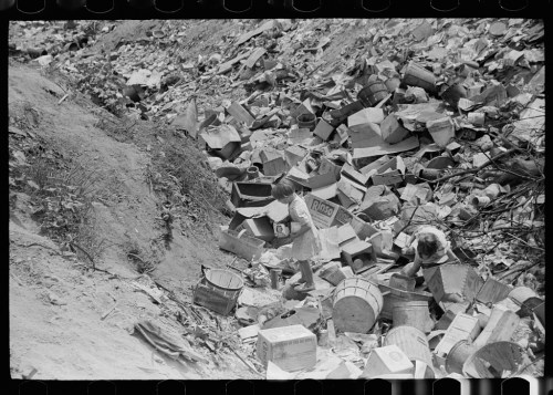 Two individuals sifting through a large pile of discarded boxes and debris in a waste area.