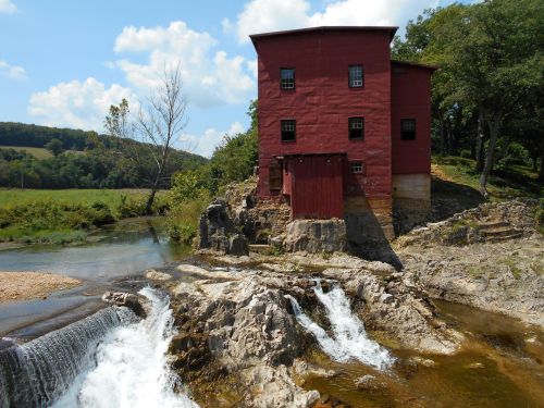 A red historic building beside a flowing river and waterfall, surrounded by greenery and blue skies.