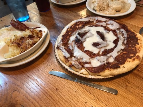 A large cinnamon roll pancake drizzled with icing, served alongside hash browns and a plate of scrambled eggs.