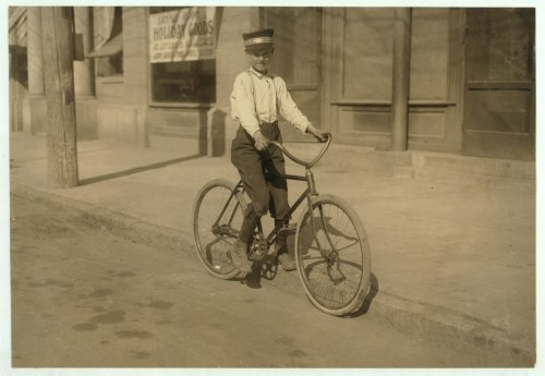 A young boy in a cap poses on a vintage bicycle in front of a store, with a dirt road and buildings in the background.