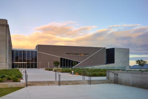 Modern building with angular design, large glass windows, and a landscaped entrance under a colorful sunset sky.