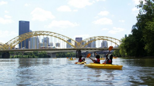 A group of kayakers paddles on a river with a city skyline and a yellow arch bridge in the background.