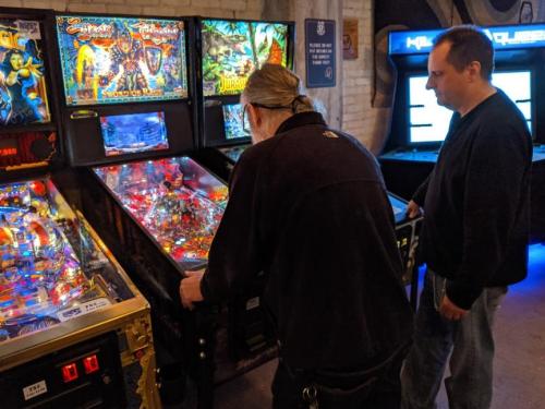Two men play pinball at an arcade, focused on the game with colorful machines in the background.