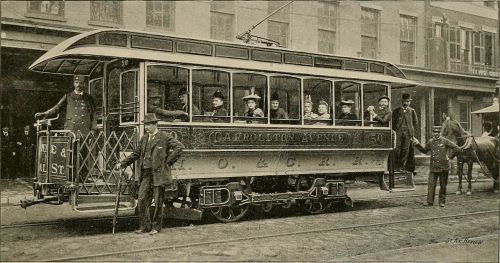 A vintage streetcar with passengers, parked on a city street, surrounded by buildings and a horse-drawn carriage.