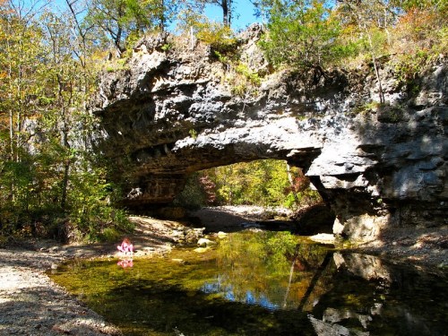 A natural stone arch over a clear stream, surrounded by trees in a vibrant autumn landscape.