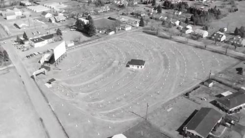 Aerial view of a drive-in theater with a large screen, surrounded by a parking area and nearby houses. Black and white image.
