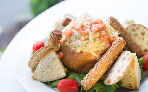 A bowl of creamy dip topped with tomatoes, surrounded by breadsticks, crackers, and cherry tomatoes on a bed of lettuce.