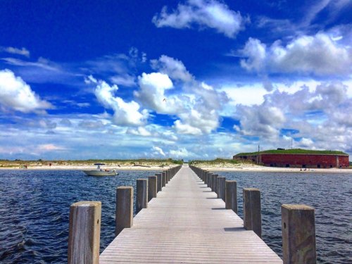 A wooden pier extends into calm waters under a bright blue sky with fluffy clouds, leading to a sandy beach.