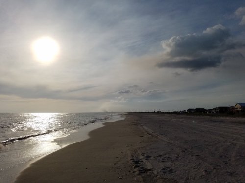 A serene beach scene at sunset, with gentle waves, soft sand, and a glowing sun in a partly cloudy sky.