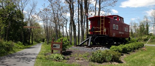 A red caboose on display near a walking path, surrounded by trees and greenery under a blue sky.