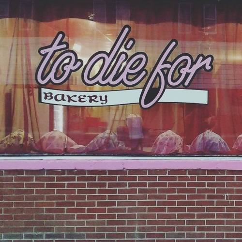 A bakery window with the sign "to die for" and a display of baked goods behind a sheer curtain.