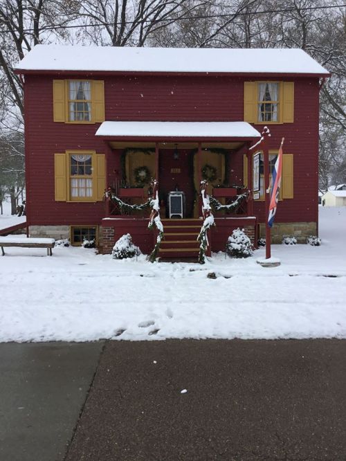 A red two-story house with yellow shutters, decorated for winter, surrounded by snow and a flag in front.