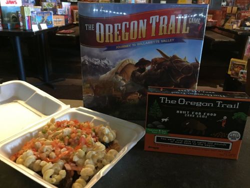 A takeout box of food topped with cheese and tomatoes, next to two Oregon Trail board games in a bookstore setting.
