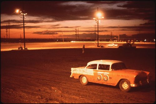A vintage race car with the number 55 parked on a dirt track at sunset, with lights illuminating the scene.
