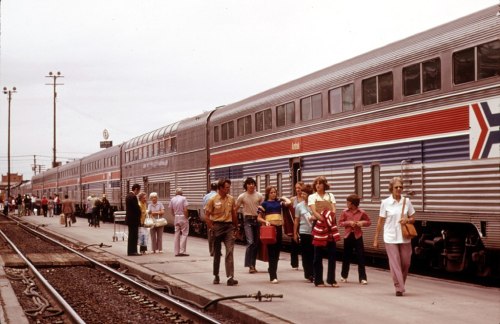 Passengers disembark from a vintage train at a station, with people waiting and walking along the platform.