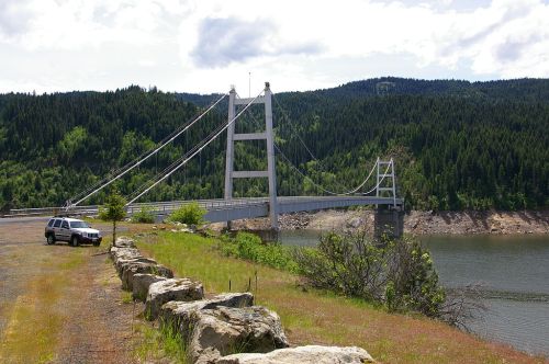 A cable-stayed bridge spans a river, surrounded by lush green hills and a parked SUV nearby.